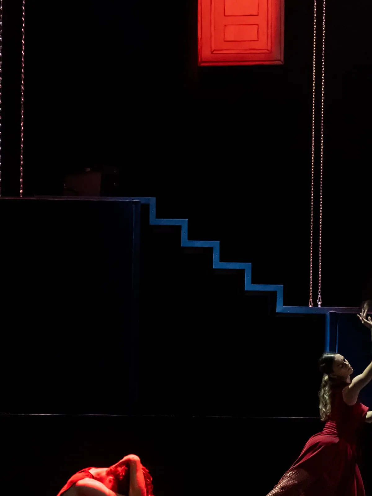 Dunkle Bühne mit rot leuchtender Tür und blauer Treppe; eine Tänzerin in Rot streckt die Arme nach oben.
